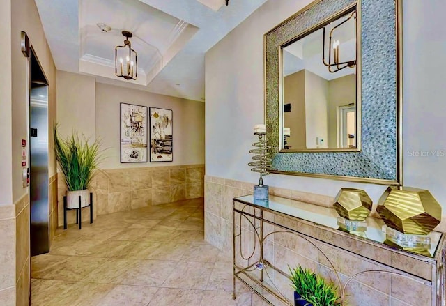 hall with a wainscoted wall, a notable chandelier, tile walls, a raised ceiling, and ornamental molding