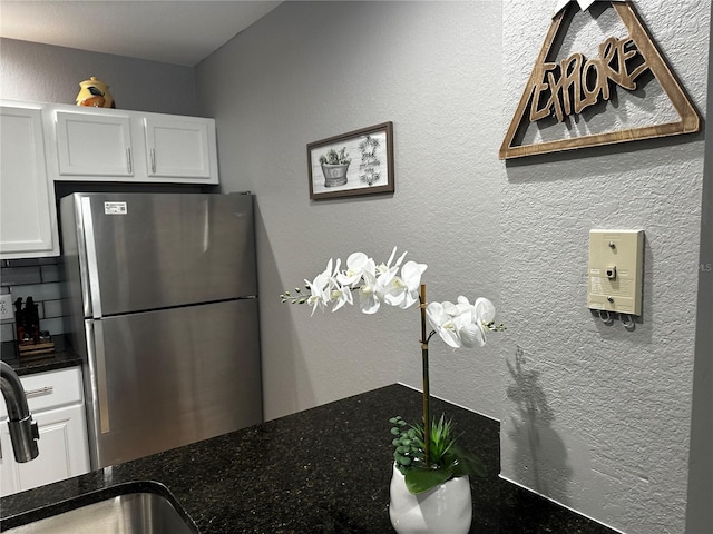 kitchen with a textured wall, dark stone countertops, freestanding refrigerator, white cabinetry, and a sink