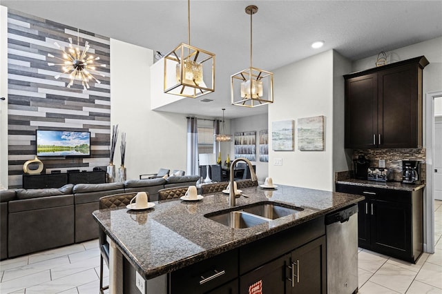 kitchen with dark stone counters, an island with sink, a sink, stainless steel dishwasher, and a notable chandelier