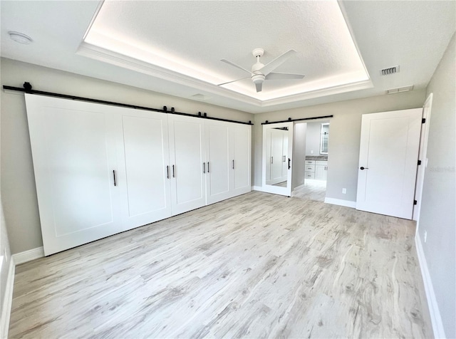 unfurnished bedroom featuring light hardwood / wood-style flooring, a textured ceiling, ceiling fan, a raised ceiling, and a barn door