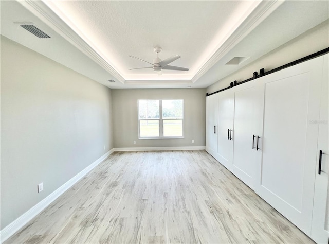 unfurnished bedroom with ceiling fan, light hardwood / wood-style flooring, a tray ceiling, and a barn door