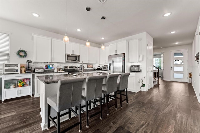 kitchen with dark stone counters, dark wood-type flooring, white cabinetry, stainless steel appliances, and a center island with sink