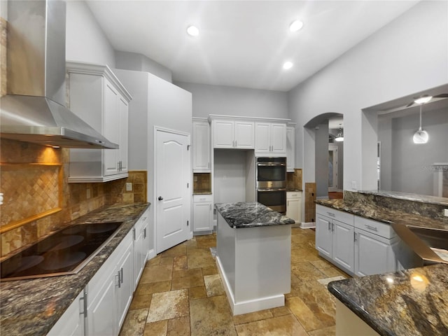 kitchen with black electric stovetop, stone tile floors, double oven, white cabinets, and wall chimney exhaust hood