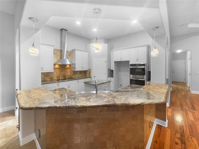 kitchen featuring white cabinets, wall chimney exhaust hood, dark stone countertops, stainless steel double oven, and pendant lighting