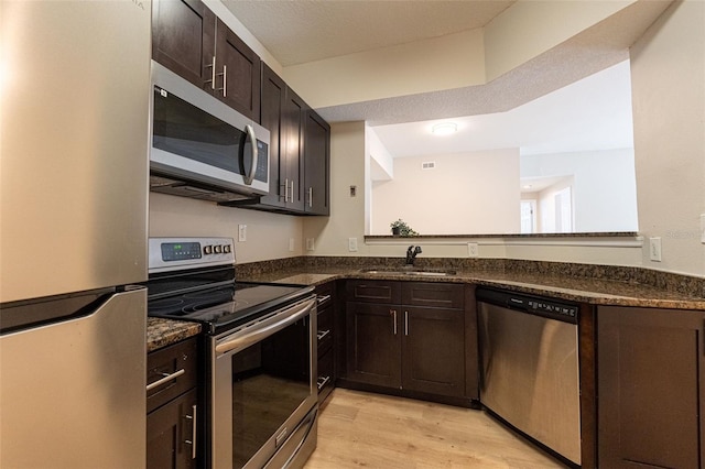 kitchen with sink, dark brown cabinets, stainless steel appliances, dark stone counters, and light wood-type flooring