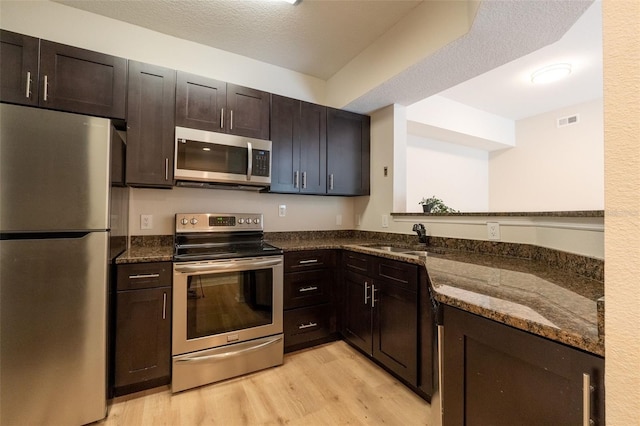 kitchen with dark brown cabinets, light hardwood / wood-style floors, dark stone counters, and appliances with stainless steel finishes