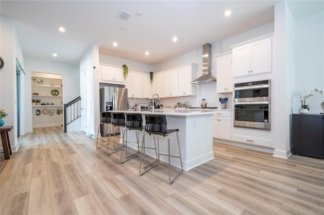 kitchen featuring appliances with stainless steel finishes, light hardwood / wood-style floors, white cabinetry, wall chimney range hood, and an island with sink