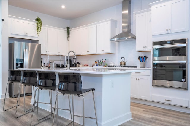 kitchen with an island with sink, light hardwood / wood-style floors, wall chimney range hood, and tasteful backsplash