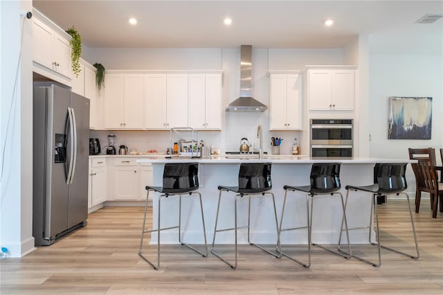 kitchen with light wood-type flooring, appliances with stainless steel finishes, a center island with sink, and wall chimney range hood