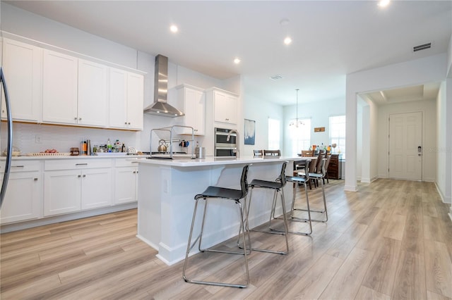 kitchen featuring light wood-type flooring, wall chimney exhaust hood, white cabinetry, and a center island with sink