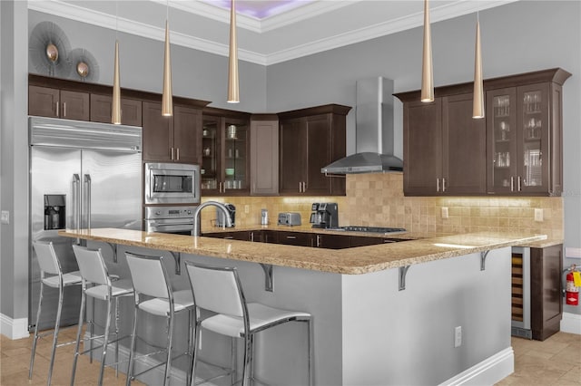 kitchen featuring kitchen peninsula, built in appliances, hanging light fixtures, wall chimney exhaust hood, and tasteful backsplash