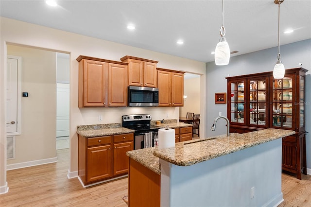 kitchen with pendant lighting, appliances with stainless steel finishes, an island with sink, light hardwood / wood-style floors, and light stone counters