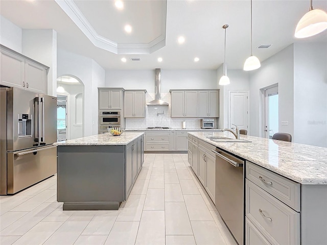 kitchen with sink, wall chimney exhaust hood, light stone counters, an island with sink, and stainless steel appliances