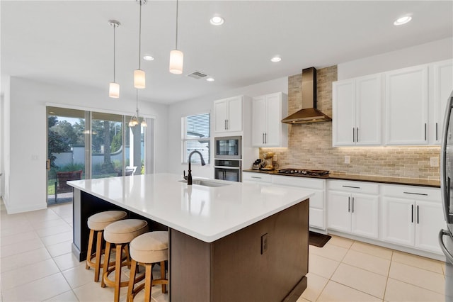 kitchen with a kitchen island with sink, stainless steel appliances, decorative light fixtures, sink, and wall chimney range hood