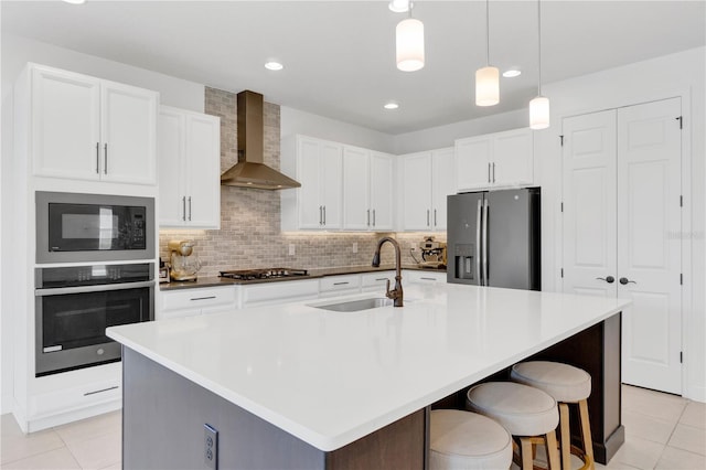 kitchen featuring a center island with sink, wall chimney exhaust hood, stainless steel appliances, sink, and white cabinetry