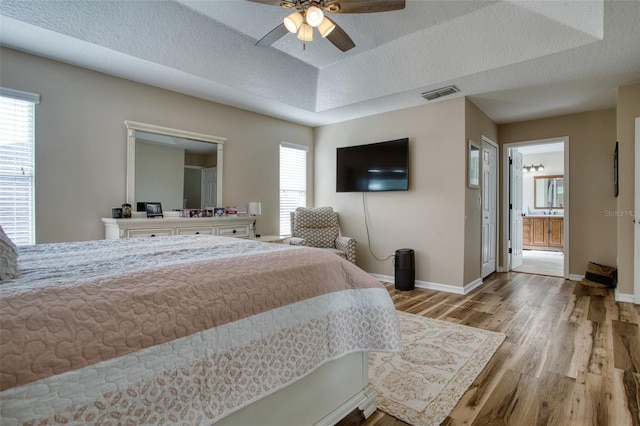 bedroom featuring ceiling fan, a raised ceiling, light hardwood / wood-style flooring, ensuite bathroom, and a textured ceiling