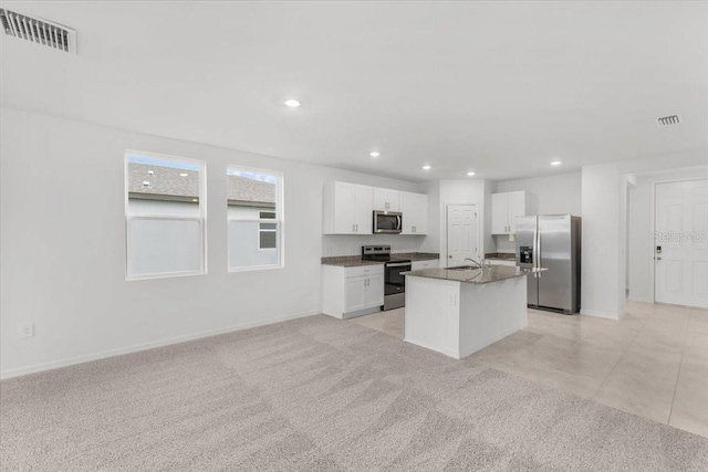 kitchen featuring a center island with sink, appliances with stainless steel finishes, white cabinetry, dark stone counters, and light colored carpet