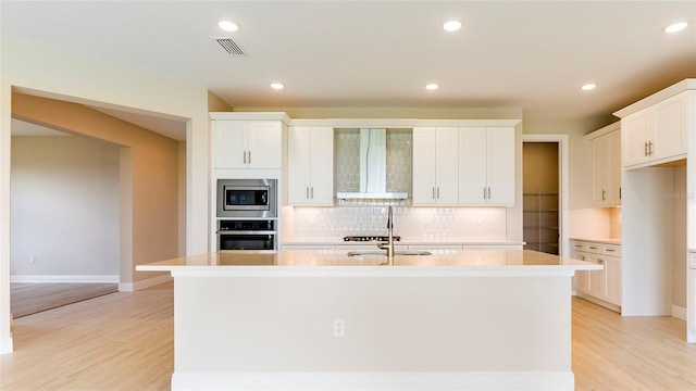 4845 Carova Way, Lakewood Ranch, FL 34211 - kitchen with stainless steel appliances, wall chimney exhaust hood, a kitchen island with sink, backsplash, and light wood-style flooring