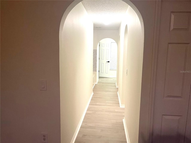 corridor with arched walkways, baseboards, light wood finished floors, and a textured ceiling