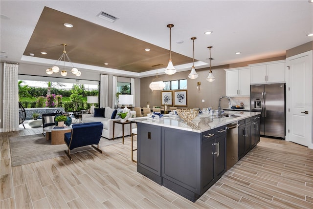 kitchen with sink, a raised ceiling, stainless steel appliances, a center island with sink, and white cabinets