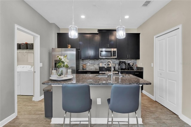 kitchen with appliances with stainless steel finishes, backsplash, washer and clothes dryer, sink, and an island with sink