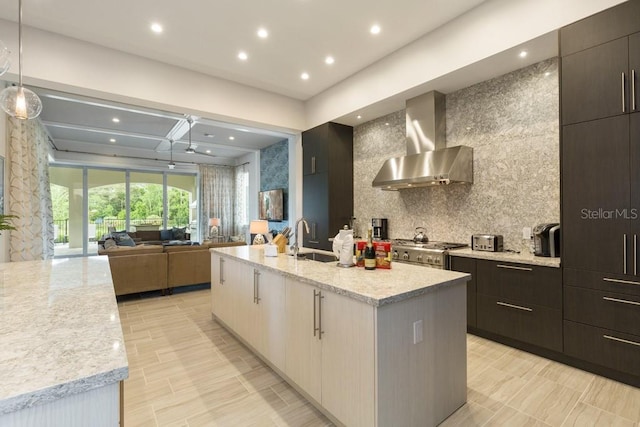 kitchen featuring decorative light fixtures, sink, light stone counters, a center island with sink, and wall chimney range hood