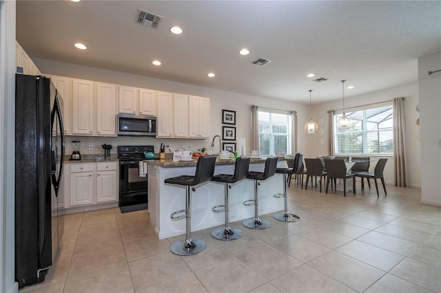 kitchen with an island with sink, black appliances, pendant lighting, white cabinets, and light tile patterned floors