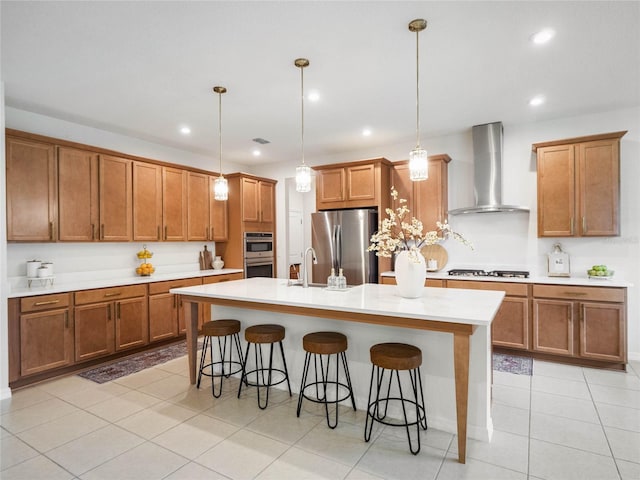 kitchen with a breakfast bar area, a center island with sink, appliances with stainless steel finishes, hanging light fixtures, and wall chimney range hood