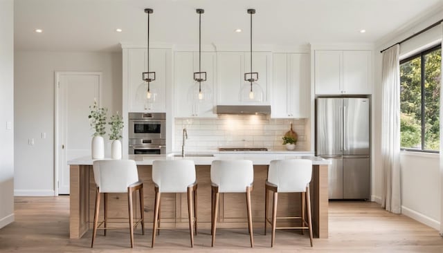 kitchen featuring light wood-type flooring, stainless steel appliances, a center island with sink, and decorative light fixtures