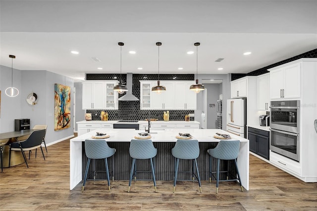 kitchen featuring high end fridge, wall chimney exhaust hood, pendant lighting, a kitchen island with sink, and double oven