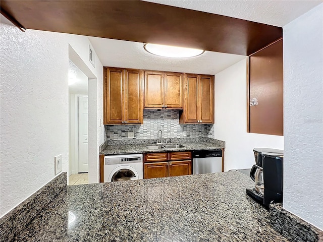 kitchen with stainless steel dishwasher, tasteful backsplash, washer / dryer, sink, and dark stone countertops