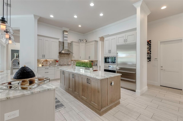 kitchen featuring built in appliances, light stone counters, a kitchen island with sink, wall chimney range hood, and decorative backsplash