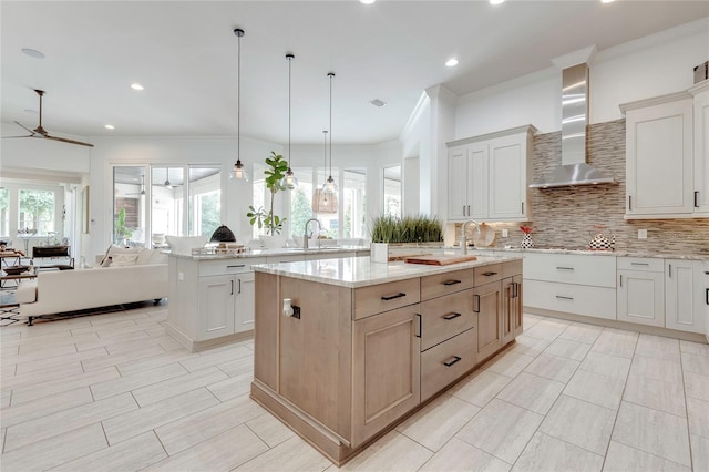 kitchen with backsplash, a ceiling fan, open floor plan, a kitchen island with sink, and wall chimney exhaust hood