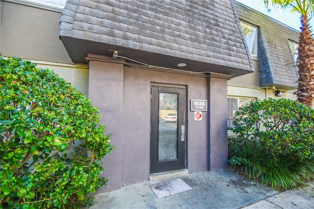 view of exterior entry with roof with shingles, mansard roof, and stucco siding