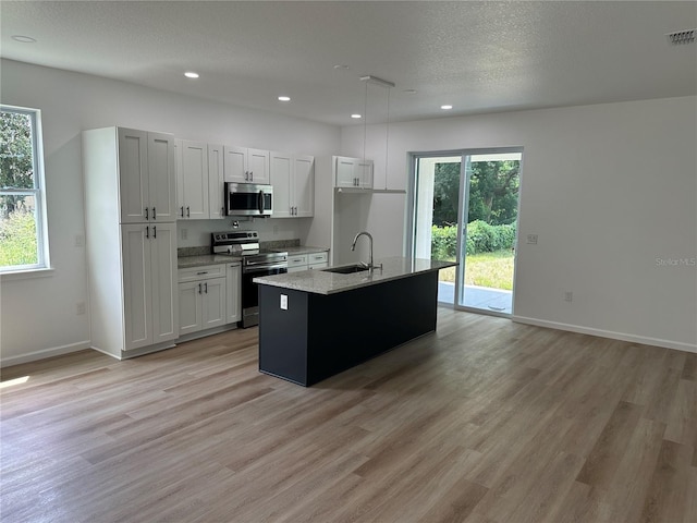 kitchen with decorative light fixtures, appliances with stainless steel finishes, white cabinets, a sink, and an island with sink