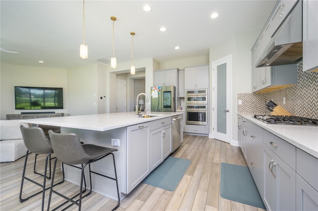 kitchen with an island with sink, pendant lighting, light hardwood / wood-style floors, exhaust hood, and appliances with stainless steel finishes