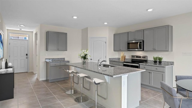 kitchen featuring appliances with stainless steel finishes, light stone counters, an island with sink, light tile patterned floors, and gray cabinets