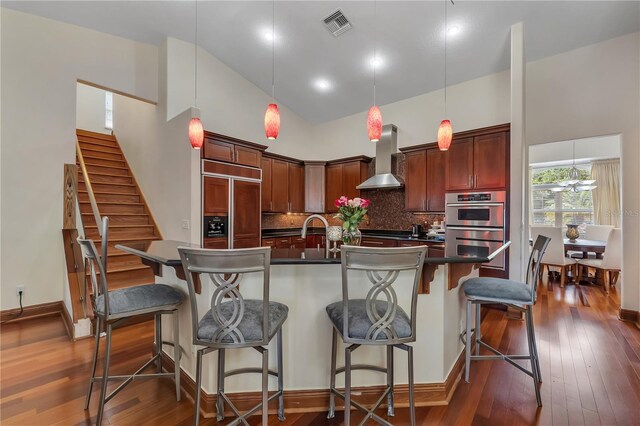 kitchen with backsplash, dark wood-type flooring, wall chimney range hood, and double oven