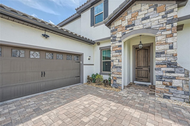 view of exterior entry with decorative driveway, stucco siding, a garage, stone siding, and a tiled roof