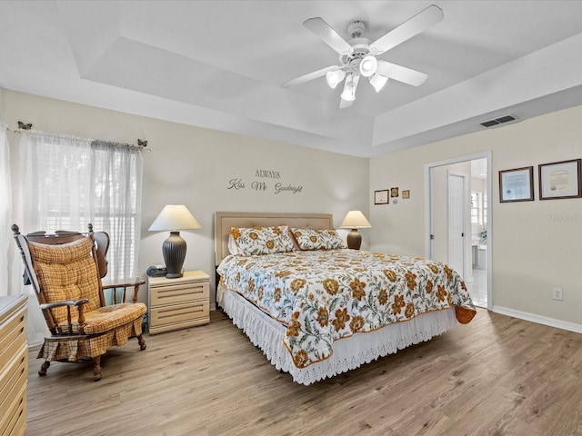 bedroom featuring connected bathroom, a tray ceiling, ceiling fan, and light wood-type flooring