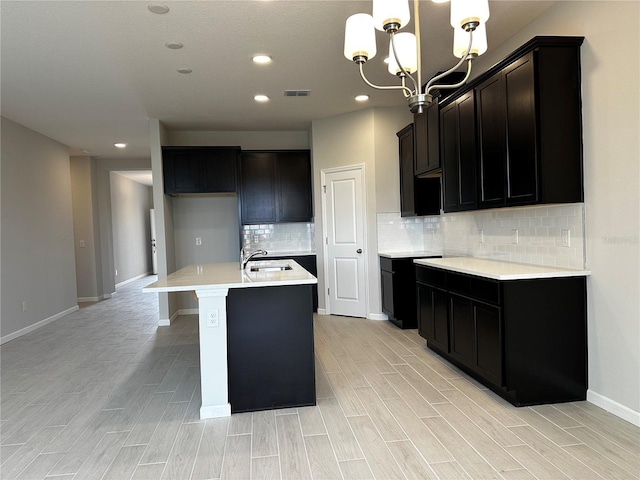 kitchen featuring tasteful backsplash, a kitchen island with sink, sink, decorative light fixtures, and a notable chandelier