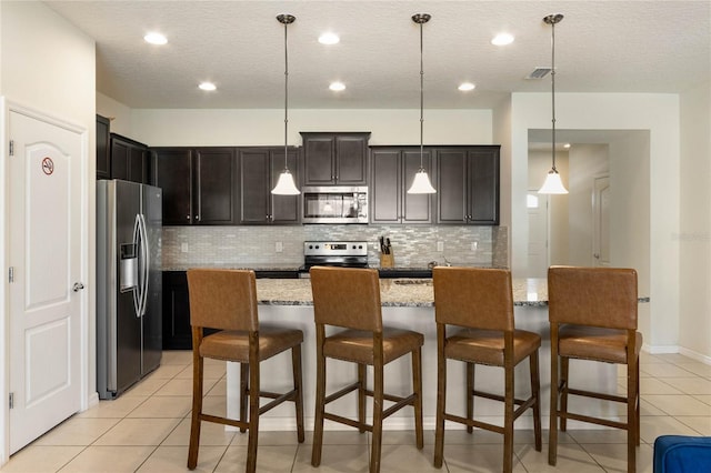 kitchen featuring light stone counters, decorative light fixtures, a center island with sink, appliances with stainless steel finishes, and a kitchen bar