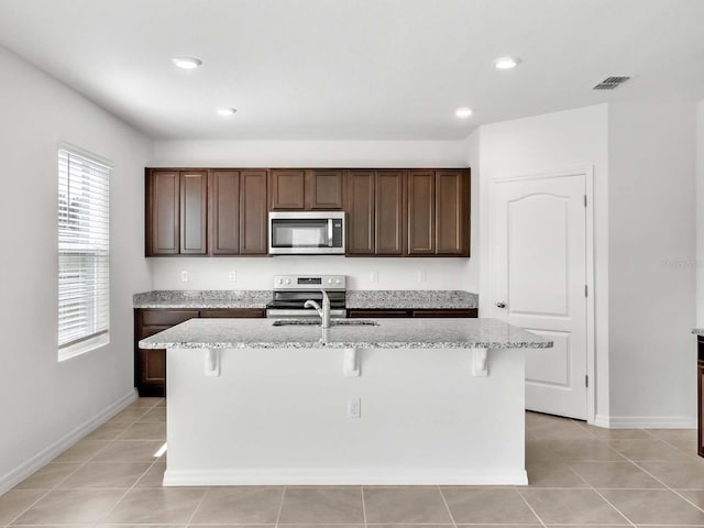 kitchen with stainless steel appliances, dark brown cabinets, a center island with sink, light stone countertops, and light tile patterned floors