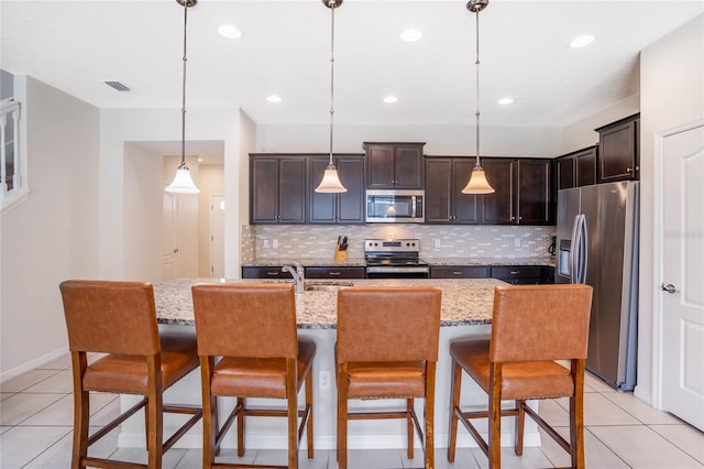 kitchen with an island with sink, light stone counters, a kitchen breakfast bar, decorative light fixtures, and stainless steel appliances