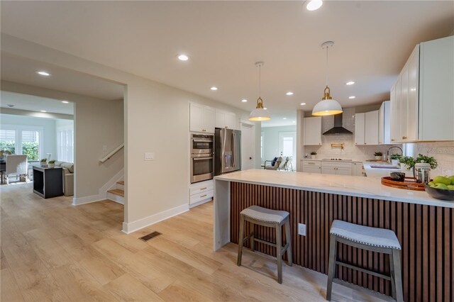 kitchen with stainless steel appliances, light countertops, hanging light fixtures, white cabinetry, and wall chimney range hood