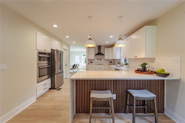 kitchen with a peninsula, a sink, light countertops, wall chimney range hood, and appliances with stainless steel finishes