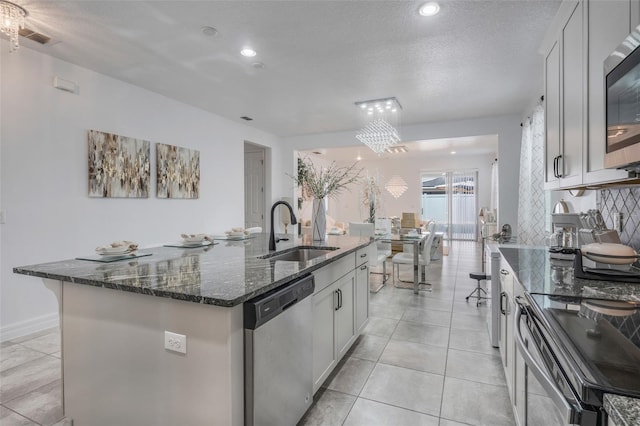 kitchen with a textured ceiling, stainless steel appliances, sink, a center island with sink, and dark stone countertops