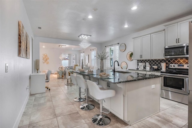 kitchen with a kitchen island with sink, dark stone counters, sink, appliances with stainless steel finishes, and tasteful backsplash