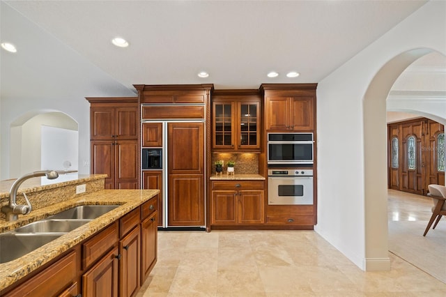 kitchen with arched walkways, paneled fridge, stainless steel double oven, a sink, and decorative backsplash