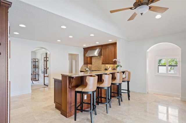 kitchen with light stone countertops, wall chimney exhaust hood, kitchen peninsula, ceiling fan, and a kitchen bar